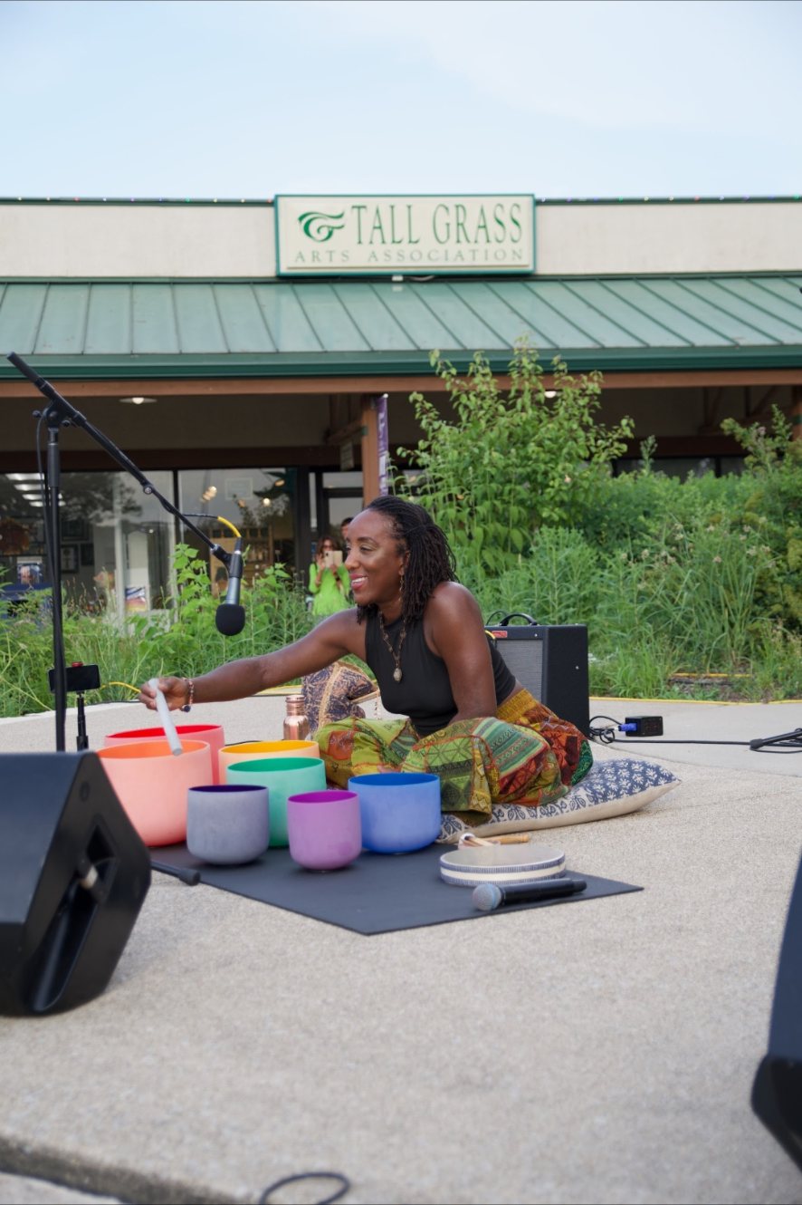 Andrea seated outdoors playing a row of crystal singing bowls in front of the Tall Grass Arts Association