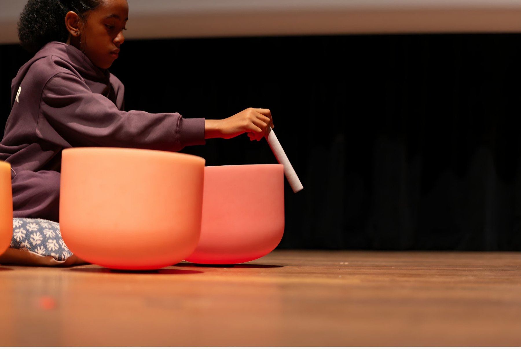 Ava Rae playing a pink crystal singing bowl on a wooden stage