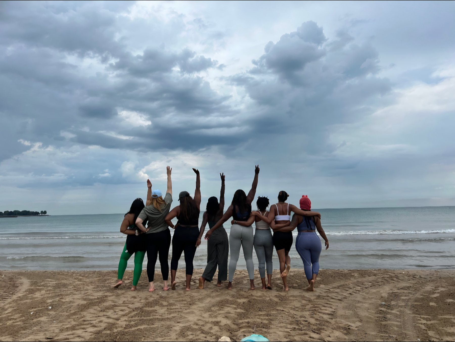 Eight women standing on a sandy shore with arms raised toward one another beneath a storm-lit sky