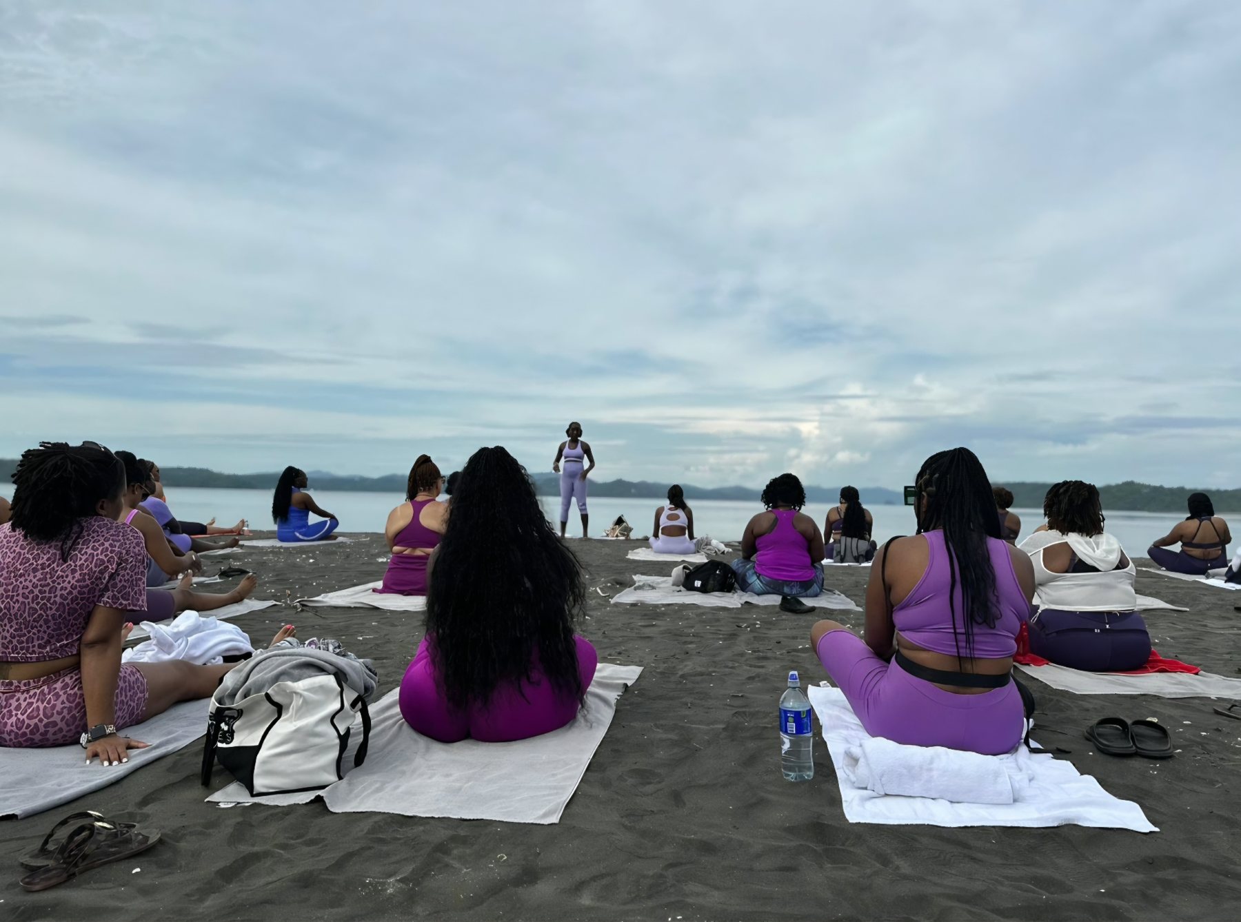 A shoreline meditation with participants seated on towels on dark sand, facing the water