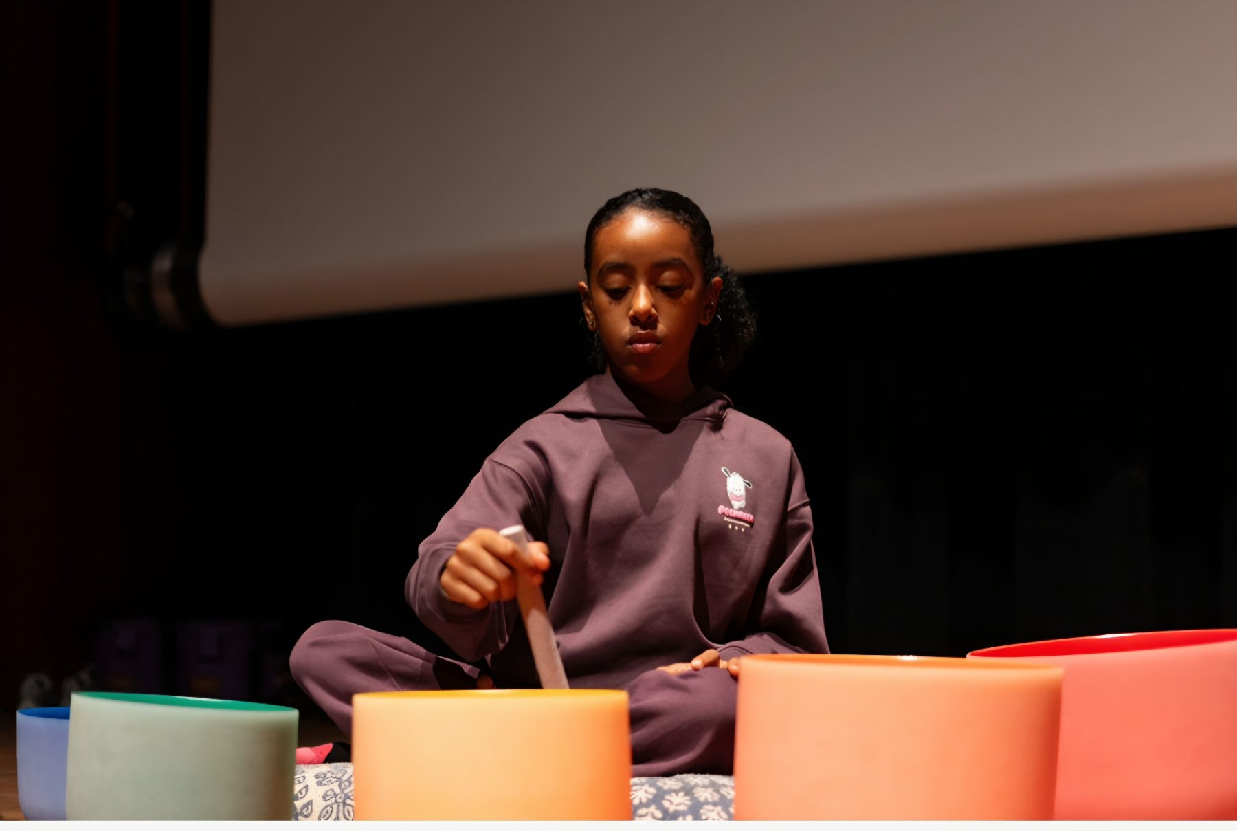 Ava Rae seated cross-legged on stage with a mallet raised over a set of crystal singing bowls