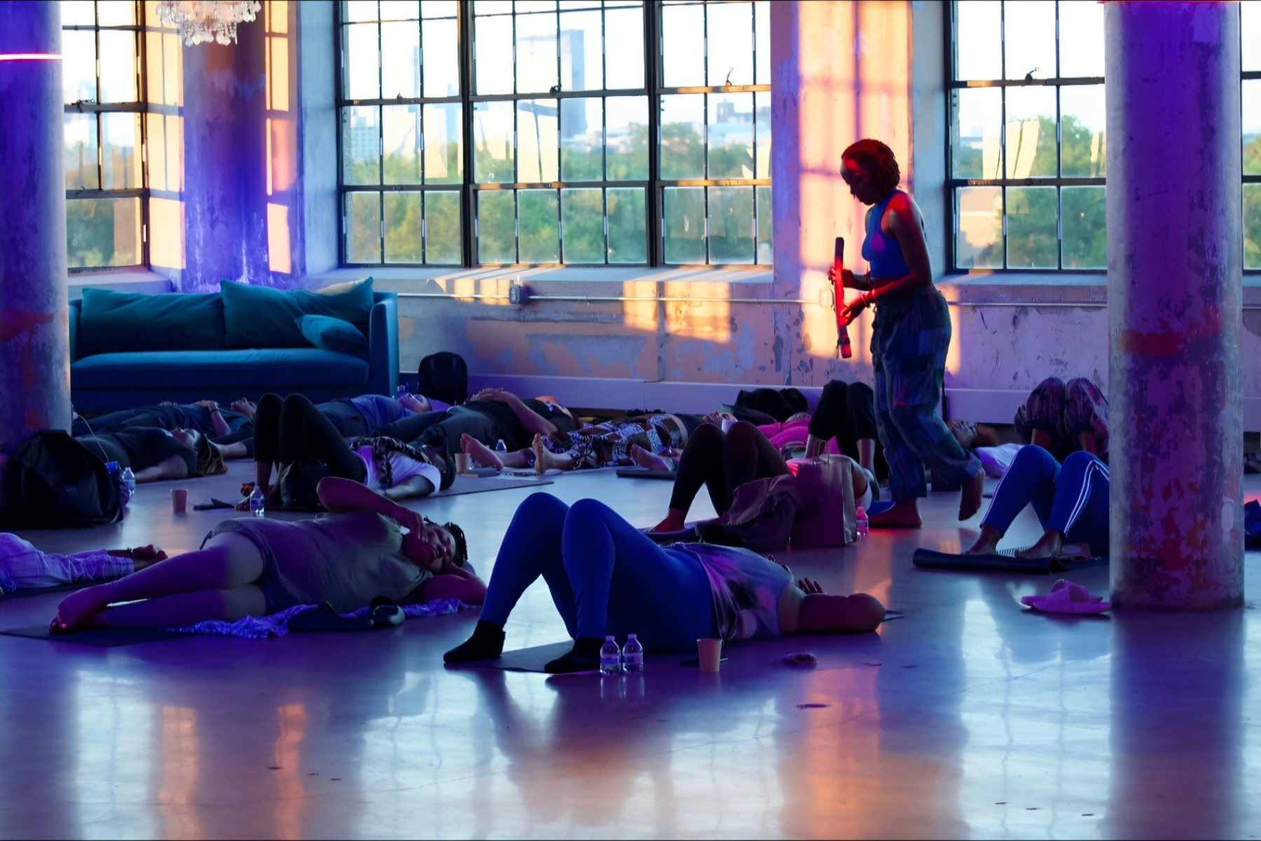 A violet-lit meditation in a loft studio, a facilitator moving between resting participants