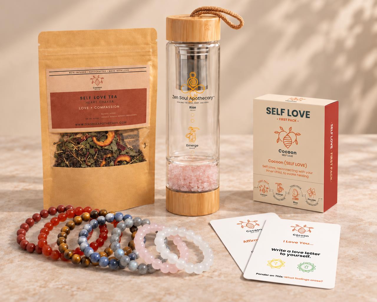 Uncluttered arrangement of Zen Soul Apothecary Self Love Tea, a glass infuser bottle, two gemstone bracelets and a small Cocoon Self Love deck with two affirmation cards